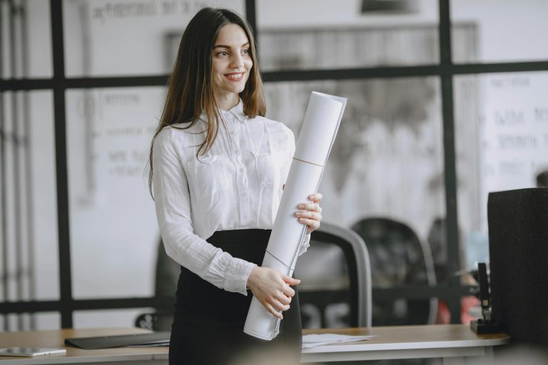 Smiling woman in office with rolled project plans, showcasing confidence and professionalism.