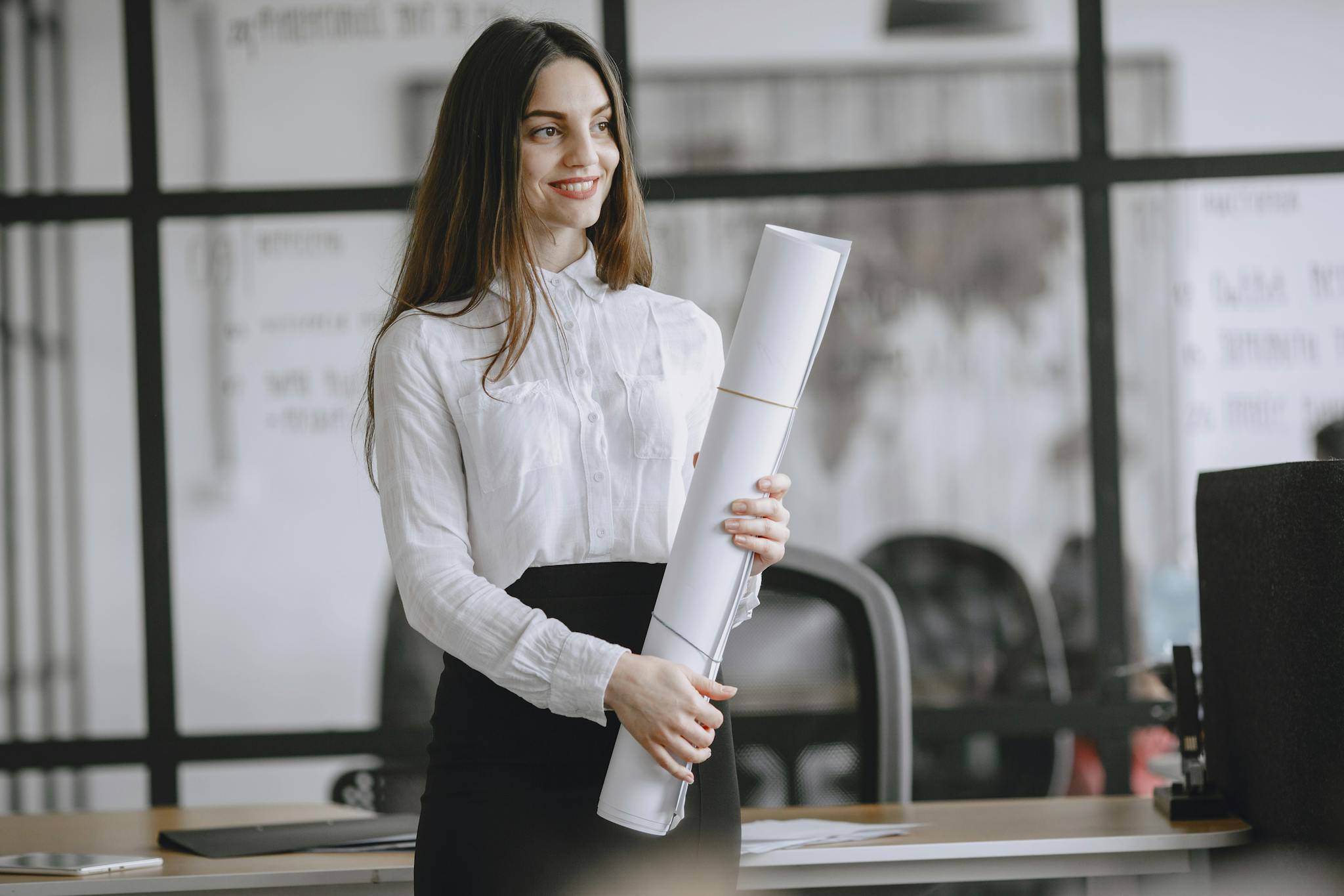 Smiling woman in office with rolled project plans, showcasing confidence and professionalism.